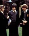 Three students in school uniforms and robes talking in a courtyard, holding books.