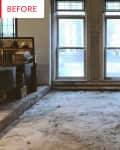 Empty living room with stained carpet, two large windows, a fireplace, and a stack of books on the left.