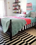 Bedroom with a black bed frame, colorful bedding, striped rug, wall shelves, and a pink typewriter art piece.