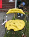Damaged yellow chair with torn cushion on grass, child playing in background.
