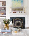 Living room with a white fireplace, abstract painting, gold candlesticks, and a glass coffee table with decor books and plants.