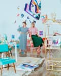 Two women in colorful outfits stand in an art-filled room with abstract wall decor and vibrant furniture.
