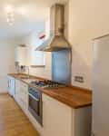 Narrow kitchen with wooden countertops, gas stove, white cabinets, bar stools, and a stainless steel fridge.