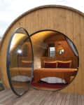 Wooden barrel-shaped cabin with a round glass door, revealing a cozy bed with orange bedding inside.