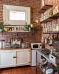 Rustic kitchen with brick walls, open shelves, white cabinets, and various kitchen items including jars, plants, and a yellow bowl.