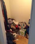 Messy closet with piles of clothes, shoes, and a green shopping bag on a wooden floor.