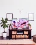 Living room with TV on dark wood stand, wicker baskets, potted plant, leather sofa, and patterned poufs.