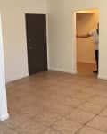 Empty kitchen with white cabinets, stainless steel sink, and beige tiled floor. Person partially visible in doorway.