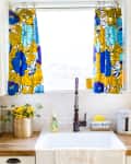 Kitchen sink with floral curtains, yellow flowers in a pot, soap dispenser, and a towel with floral embroidery.