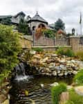 Stone house with turret, surrounded by lush greenery, overlooking a pond with a small waterfall and rock garden.