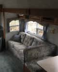 RV interior with floral sofa, two blue armchairs, small wooden table, and kitchen sink, before renovation.