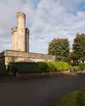 Stone tower with crenellations beside a road, surrounded by green hedges and trees under a cloudy sky.