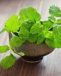 Fresh green lemon balm leaves in a rustic ceramic bowl on a wooden table.