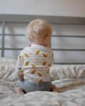 Toddler sitting on a bed with patterned sheets, wearing a bird-patterned shirt and polka dot shorts.