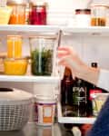 Person organizing a refrigerator with glass jars of juice, herbs, and condiments, alongside bottles and a salad spinner.