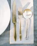 Gold knife, fork, and silver spoon on a linen napkin beside a white plate on a gray table.