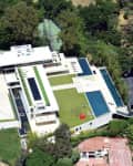 Aerial view of a modern mansion with large pools, tennis court, and landscaped gardens surrounded by trees.