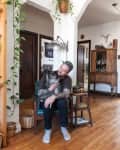 Man with tattoos sitting on a chair, holding a dog in a cozy living room with plants and wooden furniture.