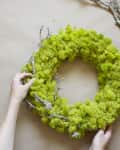 Hands holding a green moss wreath with twigs on a beige surface.