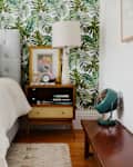 Bedroom with tropical leaf wallpaper, tufted headboard, colorful pillows, wooden nightstand, lamp, and vintage fan.