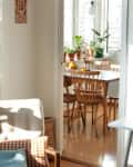 Cozy living area with armchair, colorful cushions, and a view into a sunlit dining room with wooden chairs and potted plants.