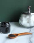 Jar of black paste, wooden spoon with powder, striped towel, and glass jar of cotton balls on marble surface.