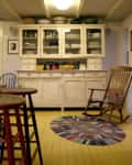 Rustic kitchen with a vintage white hutch, colorful braided rug, and wooden rocking chair on yellow floorboards.