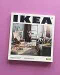 IKEA catalog on a purple surface, featuring a living room with a sofa, armchair, and colorful rug.