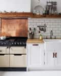 Rustic kitchen with cream AGA stove, copper backsplash, farmhouse sink, mint SMEG fridge, and wooden shelves with jars.