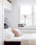 Cozy bedroom with a white bed, pink pillows, books, and plants on the windowsill, under a large window.