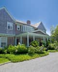Two-story house with shingle siding, covered porch, and lush garden.
