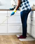 Person mopping a kitchen floor with white cabinets, wearing blue gloves and jeans, near a brick wall and countertop items.