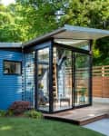Modern blue tiny house with large glass windows, wooden deck, and hot tub, surrounded by lush greenery.