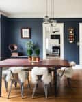 Dining room with dark blue walls, wooden table, metal chairs with white fur, and a view into a kitchen.