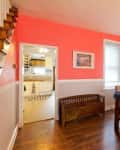 Dining room with coral walls, wooden table, beige chairs, and framed artwork, leading to a kitchen with white cabinets.