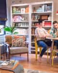 Cozy living room with bookshelves, a couple at a round table, laptop, yellow chairs, and a vase of tulips.