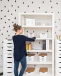 Woman organizing white storage shelves with baskets in a room with triangle-patterned wallpaper and framed art.