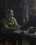 Two women sitting at a wooden table with a fruit bowl, papers, and a glass jar, in a dimly lit room with floral decor.