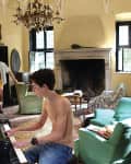 Shirtless young man playing piano in a vintage living room with green armchairs and a chandelier.