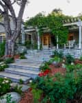 Stone house with a front porch, surrounded by lush greenery and vibrant red and white flowers.