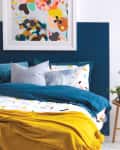 Bedroom with colorful abstract art, blue and yellow bedding, and a wooden side table with plants and a lamp.