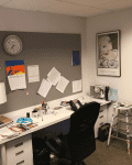 Cluttered office with a white desk, black chair, lamp, wall clock, and bulletin board with papers.
