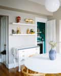 Dining room with round white table, wooden chairs, shelves with decor, and a painting on the wall.