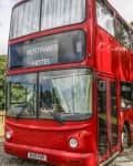 Red double-decker bus converted into Bertram's Hotel, parked on gravel with trees and wooden fence in the background.