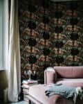 Plush mauve sofa with a gray throw, floral wallpaper, stack of books, and a silver candelabra in a cozy living room corner.