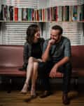 Couple sitting on a brown bench in a cozy room with bookshelves and posters on the wall.