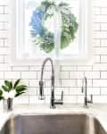 Kitchen sink with chrome faucet, white subway tiles, and a green wreath with a ribbon on the window.