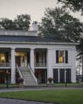 Historic white plantation-style house with large columns, black shutters, and a central staircase, surrounded by trees.