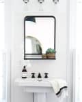 A rectangular mirror in a black frame above a white bathroom sink with black fixtures