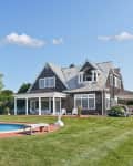 Large shingle-style house with a pool, surrounded by lush greenery and hydrangeas under a clear blue sky.